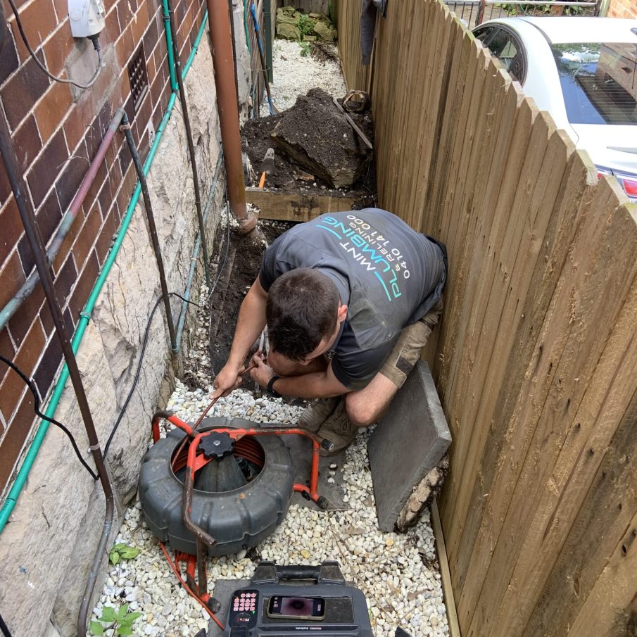 A professional plumber from Mint Plumbing & Relining working outdoors to fix a drainage system using specialised equipment, with pipes and tools visible in the background.